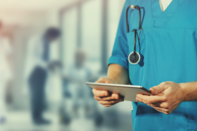 A healthcare professional in blue scrubs with a stethoscope around the neck is using a tablet, possibly managing federal health plans.