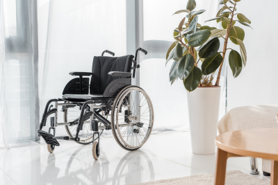 An empty wheelchair positioned next to a large potted plant in a bright room, suggesting the theme of long term disability.